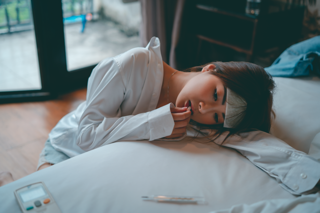 A young woman lies in bed, appearing unwell, taking medicine to alleviate symptoms.