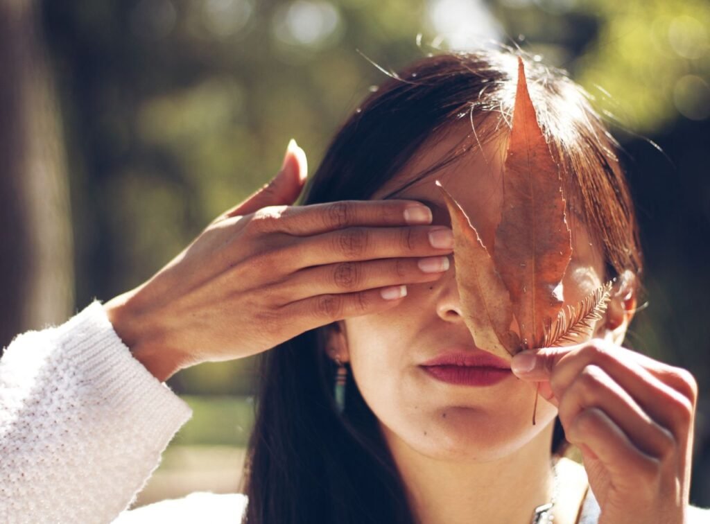 Close-up portrait of a woman outdoors, playfully covering one eye with a leaf in fall.