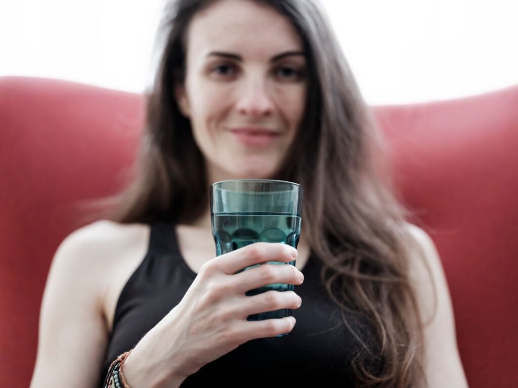 Smiling woman with long hair holding a glass of water, seated indoors with a red chair.