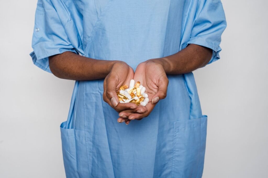 Crop faceless African American patient in blue medical robe demonstrating white and yellow pills in hands on white background in light studio