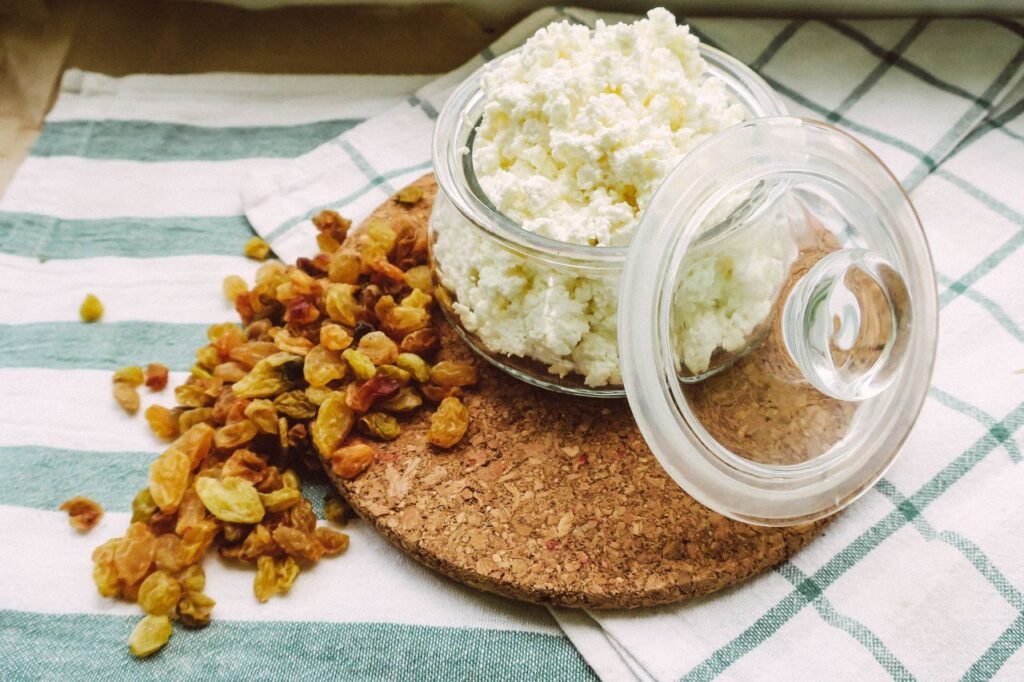 A close-up of cottage cheese in an open glass jar with scattered raisins on a cloth surface.