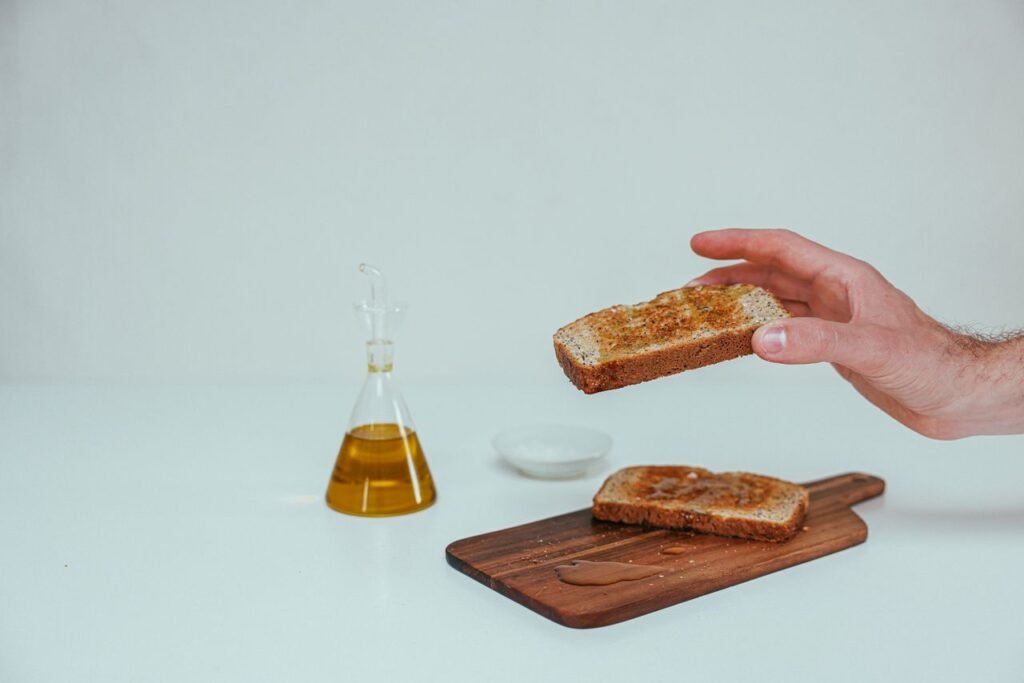 Close-up of a hand holding a slice of wheat toast, with olive oil in a bottle on a wooden board.