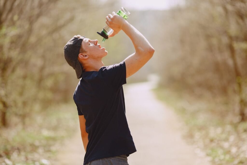 Side view of young male athlete wearing sports clothes and cap drinking water from plastic bottle on blurred background of forest during running and workout
