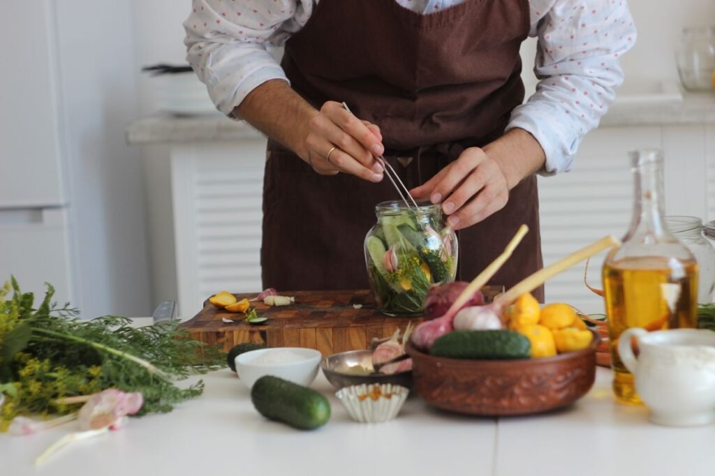 Close-up of a person pickling vegetables with tweezers in a jar, showcasing fresh ingredients on a kitchen table.